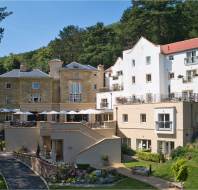 A large hotel complex with stone and white buildings, outdoor terraces with umbrellas, and landscaped gardens set against a wooded hillside.