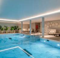 Indoor swimming pool with clear blue water, surrounded by potted plants, striped lounge chairs, and modern wall decor under recessed ceiling lights.