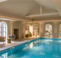 Indoor swimming pool with turquoise tiles, wicker chairs, and arched windows letting in natural light; calm and neatly arranged space.