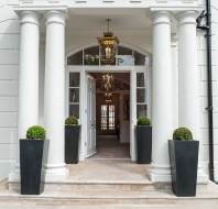 A white building entrance with four columns, large windows, double doors, lantern lights, and four tall planters with round shrubs.