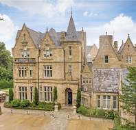 Large stone mansion with multiple gables, arched windows, and a central tower, surrounded by trees and a patio area with tables and umbrellas.