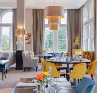 A modern restaurant dining room with large windows, elegant tables set with glassware, yellow and gray chairs, pendant lights, and floral centerpieces.