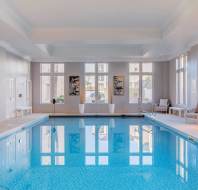 Indoor swimming pool with large windows, lounge chairs, and natural light reflecting off the water in a modern, clean setting.