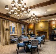 Elegant room with wooden paneling, chandeliers, blue upholstered chairs, and small tables, featuring large windows with drapes and a decorative fireplace.