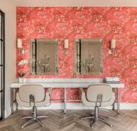 Two beige salon chairs face a white counter with mirrors, set against a bright red floral wallpapered wall in a modern, well-lit room.