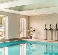 Indoor swimming pool with turquoise tiles, large windows, a potted plant, and three decorative statues on stands against a patterned wall.