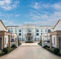 A wide driveway leads to a modern, white three-story building with columns, flanked by symmetrical single-story structures, under a blue sky with scattered clouds.