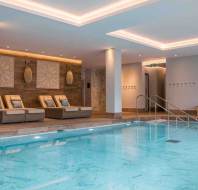 Indoor swimming pool with clear water, surrounded by lounge chairs and decorative wall art, featuring modern lighting and a large wall clock.