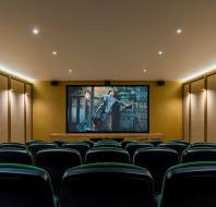 Empty home theater with green upholstered seats facing a large screen displaying a movie scene; walls are beige with recessed lighting on the ceiling.