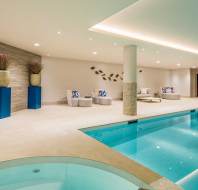 Indoor swimming pool with adjacent hot tub, modern lounge chairs with blue and white pillows, decorative plants, and neutral-toned walls illuminated by recessed lighting.