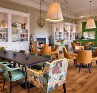 A modern dining area with wooden floors, green and yellow chairs, a long black table, and open shelves displaying decorative plates and vases.