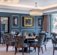 Elegant dining room with dark blue paneled walls, patterned chairs, glass tables set with dishes, large chandeliers, and a framed painting above. Large windows provide natural light.