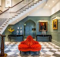 A grand foyer with a checkered marble floor, green paneled walls, a red tufted sofa, two console tables, vases, framed portraits, and a wooden staircase with white balusters.