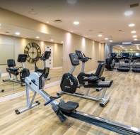 Modern gym with wooden flooring, mirrors on one wall, and various cardio equipment including treadmills, exercise bikes, and a rowing machine.