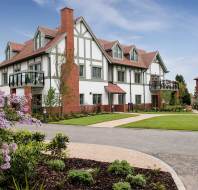A modern Tudor-style house with white walls, red brick accents, and gabled roofs, surrounded by a landscaped garden and paved driveway under a blue sky.