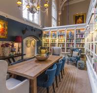 A spacious, elegant library room with tall bookshelves, a long wooden table surrounded by blue chairs, armchairs, chandeliers, and large framed portraits on the walls.