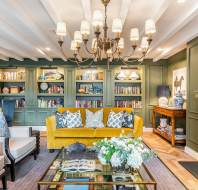 A living room with green built-in bookshelves, a yellow sofa, armchairs, a glass coffee table with flowers, a chandelier, and a fireplace.