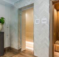A hallway features a glass door leading to a steam room and an open door to a wooden sauna, with signs labeling each. A clock and potted plant are on the left.