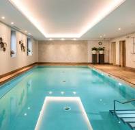 Indoor swimming pool with wooden deck, modern lighting, and wall art accents; metal handrail leads into clear blue water.
