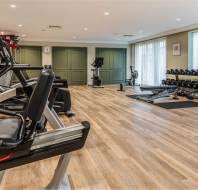 Modern gym with wooden flooring, featuring cardio machines, a row of dumbbells, weight benches, and other fitness equipment arranged neatly along the wall.