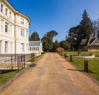 A paved driveway runs between a large white building on the left and a lawn with a round fountain and small house on the right, under a clear blue sky.