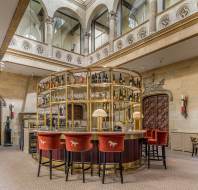 A circular bar with red velvet stools is set in a grand room with stone walls, arched windows, and ornate architectural details. Bottles line the shelves around the bar.