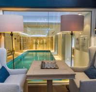 Two upholstered chairs and a chessboard table are positioned by a window overlooking an indoor swimming pool, with lamps on both sides.