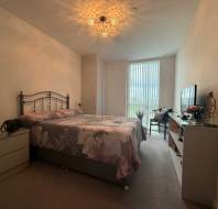 A neatly made bed with floral bedding is centered in a bedroom with minimalist decor, large window with vertical blinds, and a TV on a dresser opposite the bed.