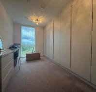A narrow, carpeted room with built-in wardrobes on one side, a dresser with a mirror, a small desk by the window, and a wicker bench below the window.
