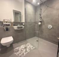Modern bathroom with a wall-mounted toilet, small sink, glass-enclosed shower, gray tiled walls, and a gray and white patterned floor mat.