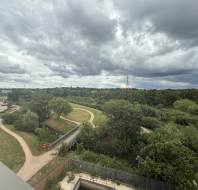 A cloudy sky over a green landscape with trees, winding paths, a bridge, and residential buildings on the left side.