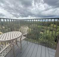 A small balcony with a metal table and two chairs overlooks a green landscape under a cloudy sky.