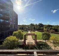 A modern apartment building overlooks a landscaped park with green lawns, walking paths, trees, benches, and a wooden pergola under a clear, sunny sky.