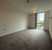 Empty, carpeted room with white walls, ceiling light, wall shelf, electrical outlets, and a large window door overlooking a building and greenery outside.