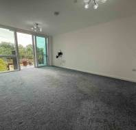 Empty living room with gray carpet, white walls, ceiling lights, and large glass doors leading to a balcony with a green outdoor view.