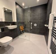 Accessible bathroom with a wall-mounted sink and toilet, walk-in shower with a folding seat and grab bars, black tiled walls, and a heated towel rail.