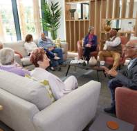 A group of elderly people sit and chat in a modern, well-lit lounge area with sofas, armchairs, and a coffee table.