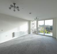 Empty, unfurnished living room with grey carpet, white walls, two ceiling lights, a radiator, and large windows with a view of a balcony and greenery outside.