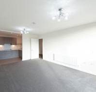 Unfurnished, bright living room with grey carpet, cream walls, ceiling lights, and an open doorway leading to a modern kitchen with grey cabinets.