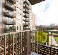 View from a balcony overlooking modern apartment buildings, a landscaped courtyard, and a green lawn with trees in the background.