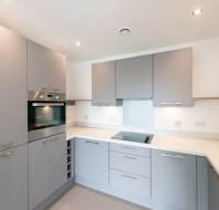 Modern kitchen with light gray cabinets, integrated oven, electric cooktop, under-cabinet lighting, and a stainless steel sink on a white countertop.