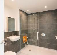 Modern accessible bathroom with dark tile walls, walk-in shower, foldable orange shower seat, grab bars, wall-mounted sink, and overhead lighting.