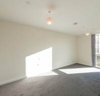 Empty, unfurnished room with beige walls, grey carpet, two ceiling lights, and a large window letting in natural light.