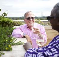 Two older adults sit outdoors, smiling and holding glasses of white wine, surrounded by greenery on a sunny day.