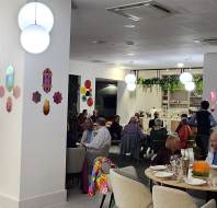 A group of people are seated at tables in a brightly lit restaurant with modern decor, wall plants, and hanging lights.