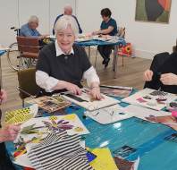 A group of older adults sit around tables covered with art supplies, making collages and crafts in a well-lit room.