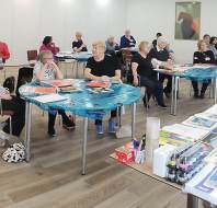 A group of adults sits at tables covered with art supplies in a classroom, listening to an instructor standing at the front near a table filled with art materials.