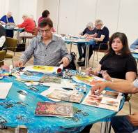 A group of adults sit at tables covered with craft supplies, magazines, and art materials, engaging in an arts and crafts activity in a bright room.