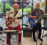 Two older men play electric guitars and sing into microphones indoors, with music stands and amplifiers nearby; large windows reveal greenery outside.
