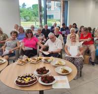 A group of older adults sits together in a bright room, smiling at the camera, with plates of assorted food and desserts on tables in the foreground.
